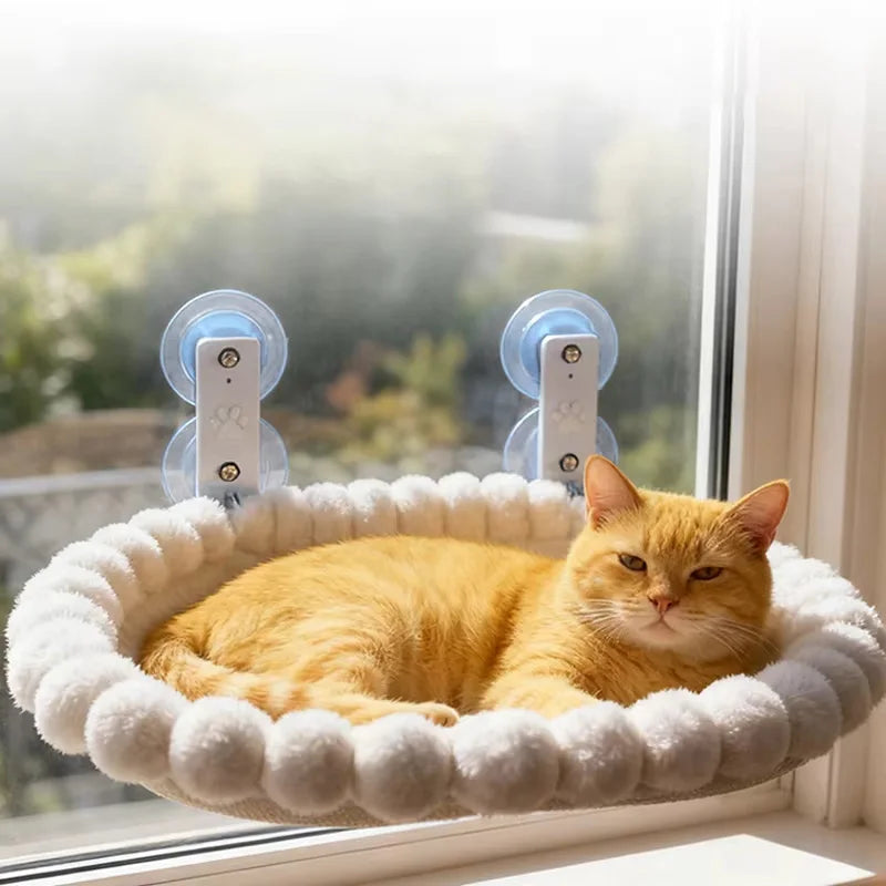 Cream pom-pom trim cat window hammock held on glass by two large suction cups with a ginger cat resting inside