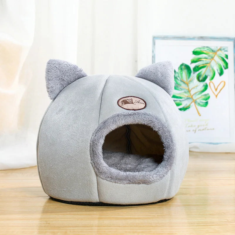 Light grey cat cave bed on a wooden floor beside framed botanical artwork